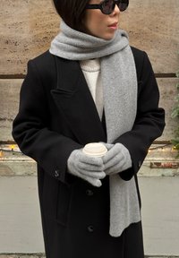 Sort uldfrakke, lysegråt ribstrikket tørklæde, grå strikkede handsker og en engangskop holdt i begge hænder mod en tekstureret væg.