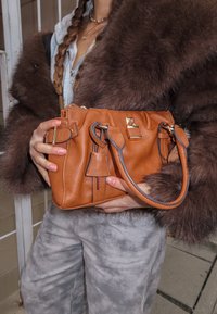 Beżowa skórzana torebka z podwójnymi uchwytami, wyposażona w złote elementy metalowe i subtelne marszczenia. Dodatkowe akcenty to ozdobne metki i gładka faktura.