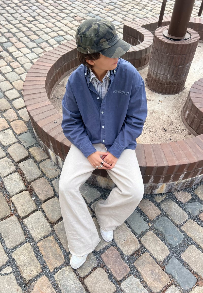 Jeune personne portant une casquette camouflage, une veste bleue et un pantalon beige large, assise sur un banc circulaire en briques sur un pavement en pavés.