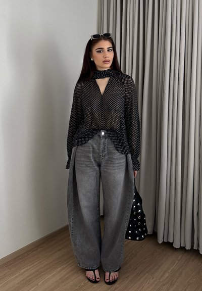 Mujer con cabello largo y oscuro que lleva una blusa negra transparente con lunares, jeans grises de tiro alto y corte ancho, sandalias negras, gafas de sol en la cabeza, y sostiene un bolso con tachuelas.