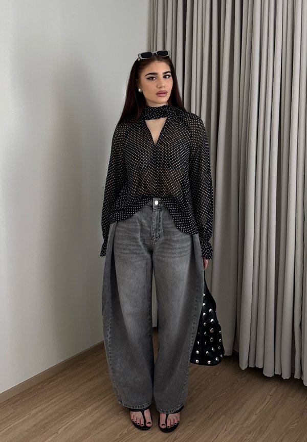 Femme aux longs cheveux foncés portant un chemisier noir transparent à pois, un jean gris large taille haute, des sandales noires, des lunettes de soleil sur la tête et tenant un sac clouté.