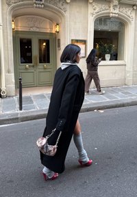 Kvinne som går på gaten iført svart kåpe, grå leggvarmere, røde hæler, holder rutete håndveske, med bygningsinngang og en annen person i bakgrunnen.