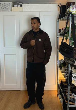 Hombre con bigote y pendientes, que lleva una chaqueta marrón y pantalones negros, está de pie en una habitación con armarios blancos y un zapatero.