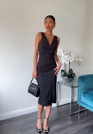 Vestido negro sin mangas con escote pronunciado, silueta ajustada y largo a la rodilla. Combinado con un pequeño bolso negro y tacones con acentos de joyas.