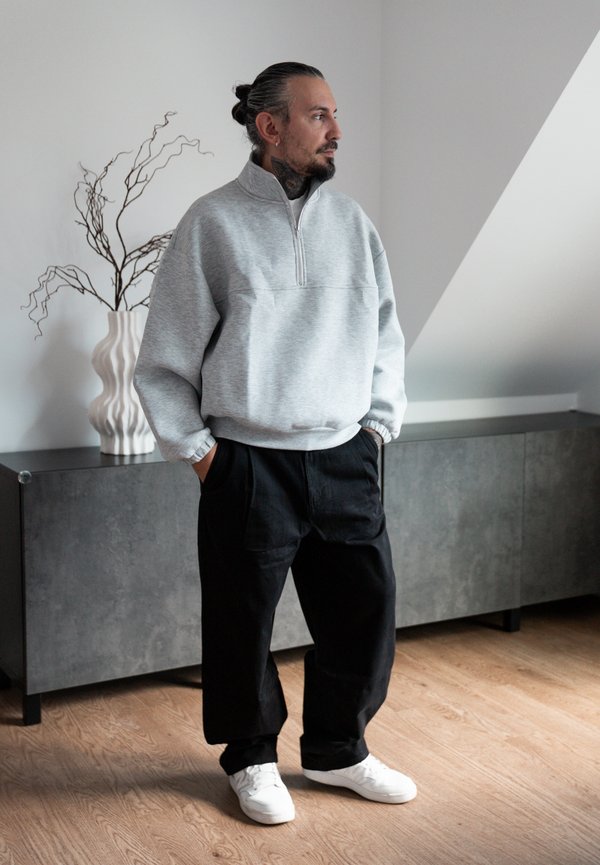 Un homme portant un sweat-shirt gris à fermeture éclair courte, un pantalon noir et des baskets blanches se tient debout, les mains dans les poches, à côté d’un meuble.