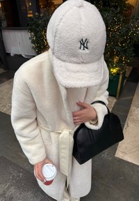 Biely plyšový župan s opaskom, nosený cez svetlý top. Krémová fleecová čiapka s čiernym logom sprevádza čiernu kabelku a šálku kávy.