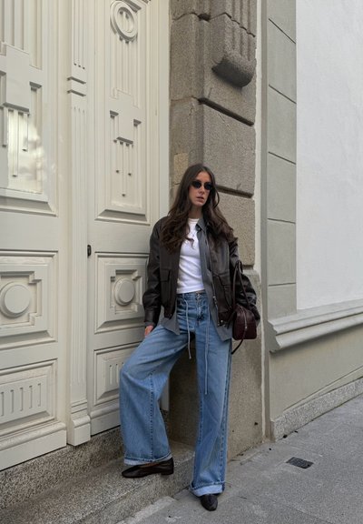 Mujer con chaqueta de cuero negra, camiseta blanca, vaqueros holgados azules y gafas de sol, apoyada contra un pilar de piedra junto a una gran puerta blanca ornamentada.