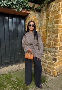 Femme portant une veste à chevrons surdimensionnée, un pantalon noir à jambes larges, des lunettes de soleil, et tenant un petit sac à main marron, debout près d'un mur en pierre et d'une porte noire.