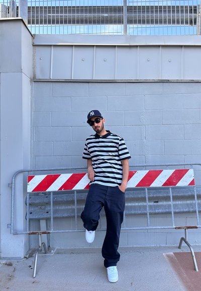 Camisa de polo de rayas blanco y negro de manga corta, pantalones oscuros, zapatillas blancas, gorra negra y gafas de sol, posando junto a una barrera.