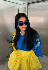 Giacca sportiva blu con zip, strisce bianche e gonna gialla. Accessori includono occhiali da sole blu e una collana di perle. Capelli texturizzati.