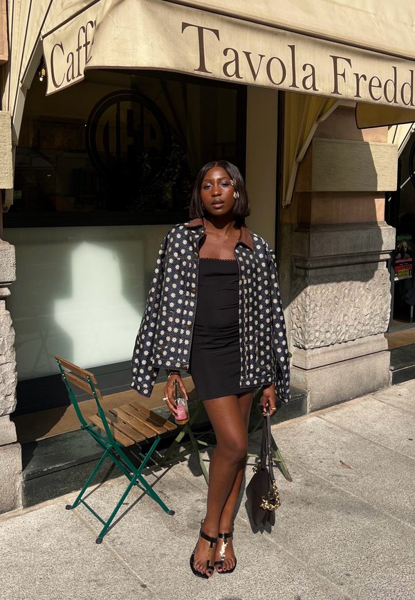 Chaqueta negra con estampado floral sobre un vestido negro. Acentos marrones en el cuello. Bolsito negro, sandalias con tirantes y detalles dorados, bebida rosa en la mano.