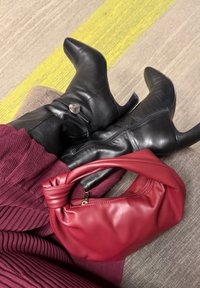 Άτομο που φοράει μαύρα δερμάτινα μποτάκια με τακούνι και λεπτομέρεια καρδιάς, μπορντό υφασμάτινη φούστα, κρατώντας μια κόκκινη δερμάτινη τσάντα με κόμπο, καθισμένο σε καναπέ.