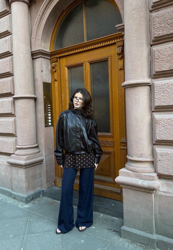 Vrouw met bril in een zwarte leren jas, polkadot blouse en donkerblauwe broek, staand bij een houten deur met stenen zuilen op een stadsstoep.