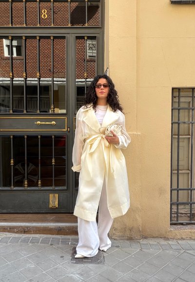 Mujer con cabello oscuro largo y rizado, que lleva gafas de sol, abrigo trench color crema, pantalones blancos y sostiene un pequeño bolso crema, de pie junto a una puerta negra y dorada.