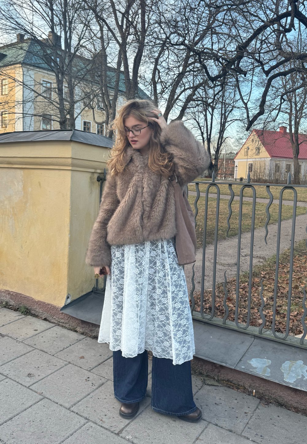 Hnědá bunda z umělé kožešiny, bílá krajková šaty, kalhoty s širokými nohavicemi z tmavého denimu a tmavé boty. Obklopena stromy a žlutou budovou.