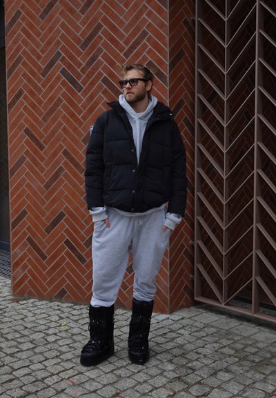 Hombre con gafas de sol, chaqueta acolchada negra, sudadera con capucha gris y pantalones deportivos, y botas negras, de pie frente a una pared de ladrillos con una puerta metálica.