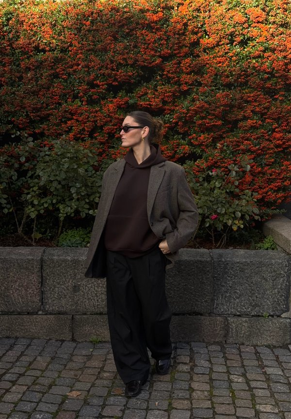 Une femme portant des lunettes de soleil, un blazer gris et des vêtements sombres se tient près d’un mur en pierre, avec des fleurs rouge orangé derrière elle.