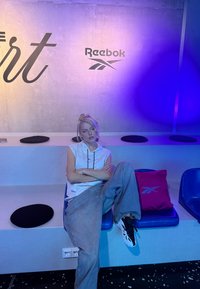 Jeune femme en tenue décontractée et sportive assise sur une chaise bleue à côté d'un coussin rose Reebok sous un éclairage violet, devant un mur en béton avec le logo Reebok.