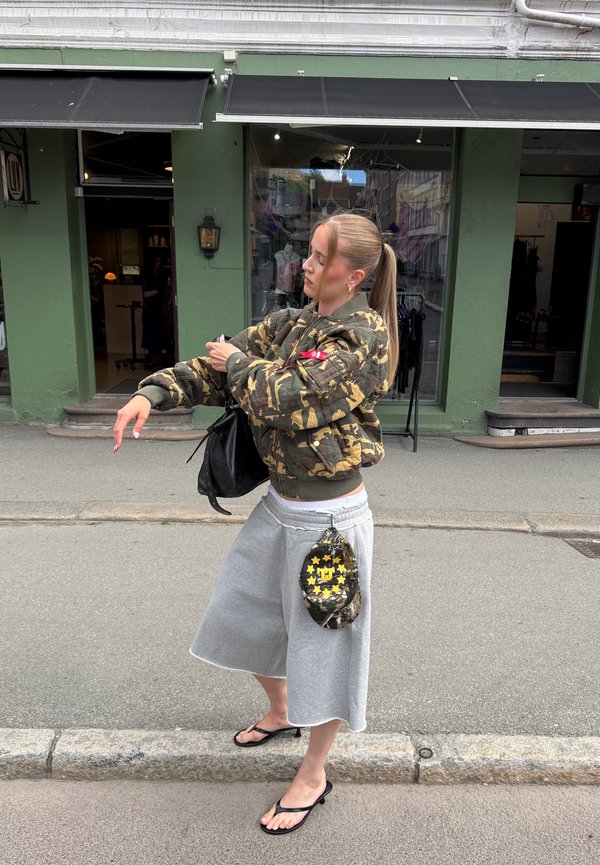 Femme en veste camouflage et short gris debout sur un trottoir en ville, portant des tongs et tenant un sac noir, avec une casquette accrochée au short.