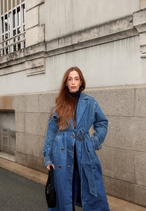 Denim trench coat with large buttons, belt, and lapels; paired with a black turtleneck. Long, straight hair cascades over shoulders.
