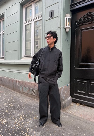 Persona vestida con un conjunto negro holgado, zapatos negros y gafas de sol está de pie en una acera de la ciudad cerca de un edificio verde pálido y una puerta negra, sosteniendo una bolsa.
