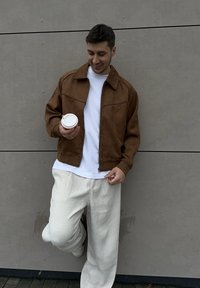 Veste en daim marron avec coutures avant, t-shirt blanc et pantalon blanc large plissé. Tenant une tasse de café, debout contre un mur gris.