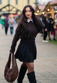 Pull marron, col blanc, jupe plissée noire, chaussettes côtelées montantes jusqu'aux genoux, et sac hobo marron, sur fond de marché en plein air.