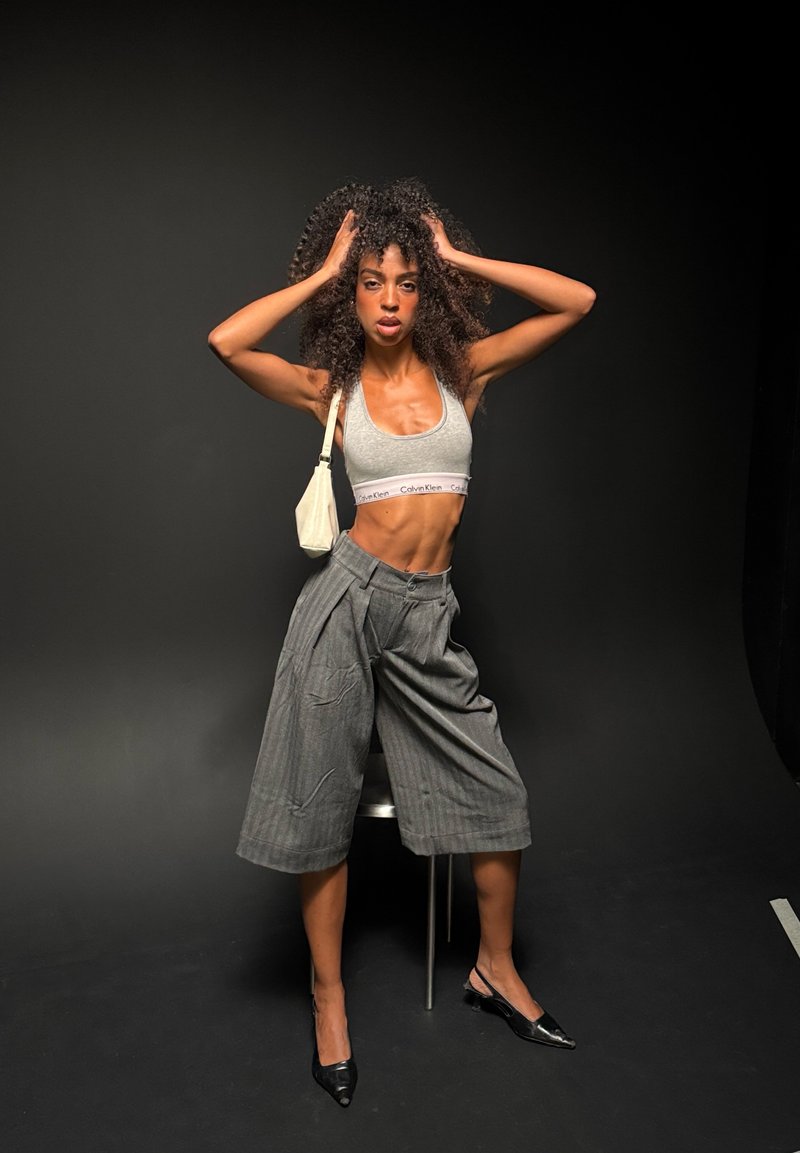 Femme aux cheveux bouclés portant un soutien-gorge de sport gris, un short large gris, des talons noirs et un petit sac à bandoulière blanc, posant les mains dans les cheveux sur un fond sombre.