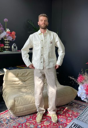 Chaqueta de denim blanca con dos bolsillos en el pecho, pantalones color crema y zapatos beige, situados contra un fondo oscuro con elementos decorativos.