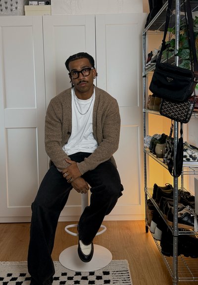 Hombre con gafas, cárdigan marrón y collar de cadena sentado en un taburete blanco junto a un zapatero con bolsas y zapatos.