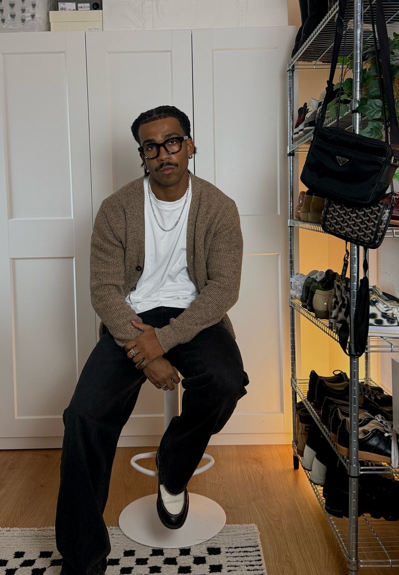 Man met bril, een bruine cardigan en een kettinghalsketting die op een witte kruk zit naast een schoenenrek met tassen en schoenen.
