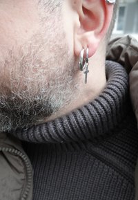 Κοντινό πλάνο του αυτιού ενός ατόμου με ασημένιο σκουλαρίκι κρίκο που φέρει ένα μικρό σταυρό, γκρίζα γένια και σκούρο ριγωτό πουλόβερ με ζιβάγκο.