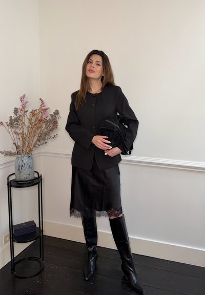 Mujer con blazer negro, falda con encaje, botas altas y bolso negro, de pie junto a una mesa lateral con un jarrón y flores secas.