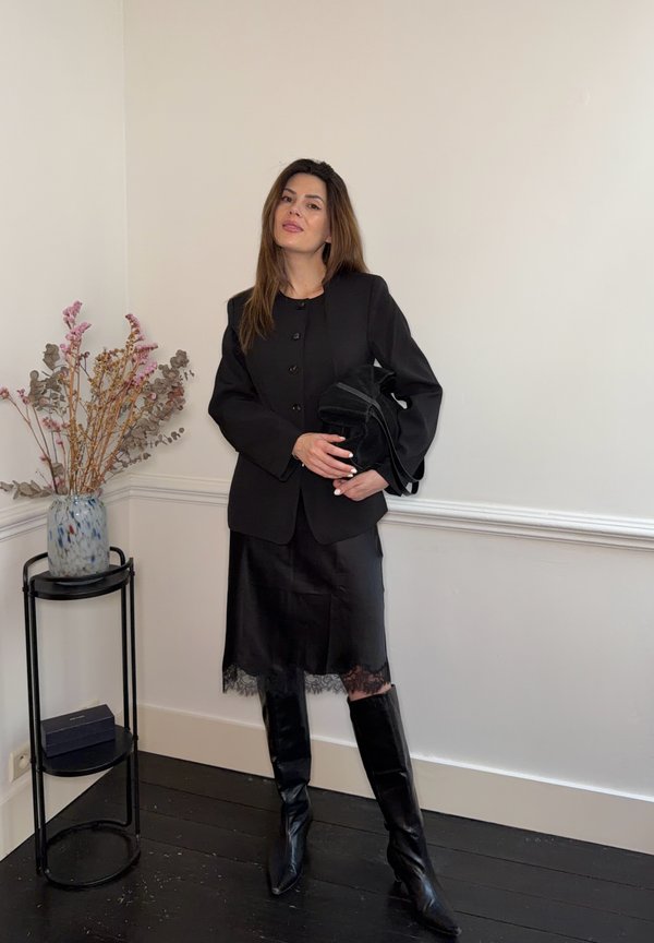 Kvinne i svart blazer, skjørt med blonder, høye støvler og en svart veske, står ved et sidebord med en vase og tørkede blomster.