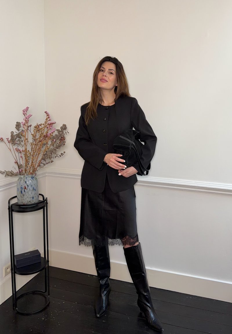 Woman in black blazer, lace-trimmed skirt, tall boots holding a black purse, standing near a side table with a vase and dried flowers.