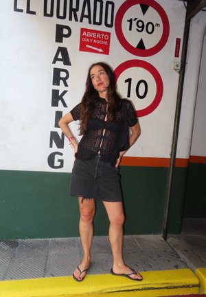 Camisa de encaje negra con un lazo frontal, combinada con una falda de mezclilla negra deshilachada y chanclas negras, con un fondo de aparcamiento.