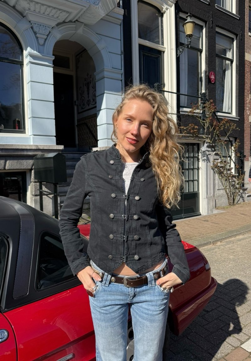 Jeune femme aux longs cheveux blonds bouclés, portant une veste noire et un jean, se tient les mains dans les poches à côté d'une voiture rouge dans une rue ensoleillée.