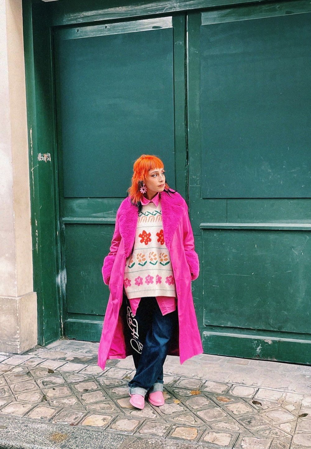 La modelo lleva un abrigo de piel sintética de color rosa brillante, un suéter de estampado floral, jeans oscuros y zapatos rosas a juego, de pie contra un fondo verde.