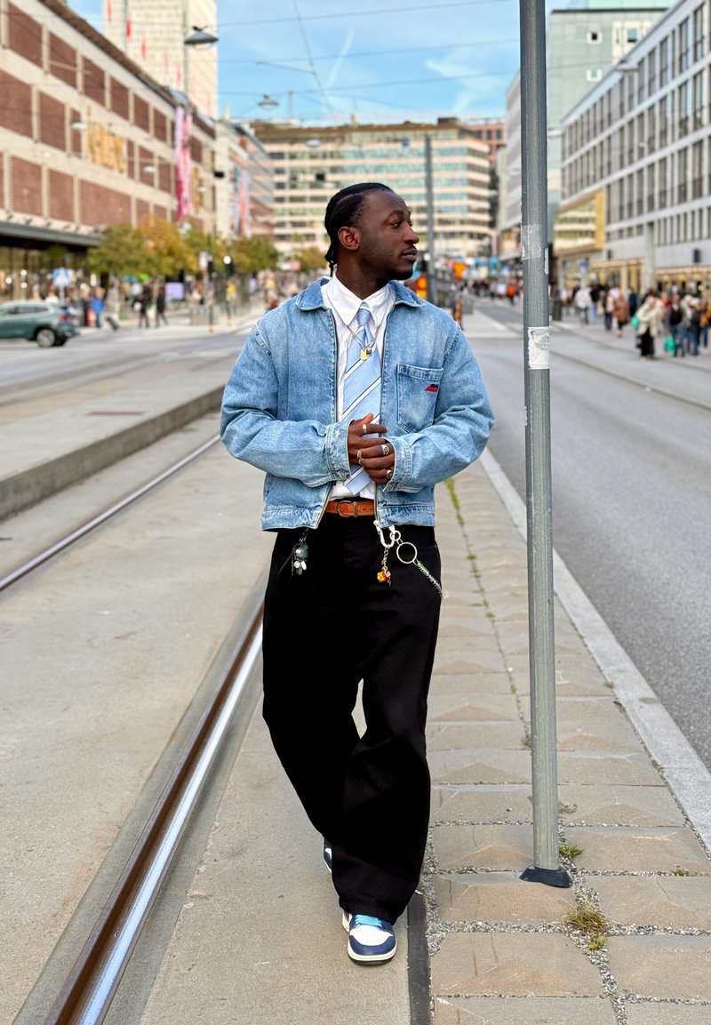 Denim jacket with a blue tie over a white shirt, black loose trousers, white and blue sneakers, and multiple accessories on a belt.
