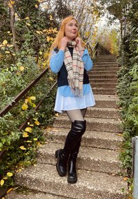 Bottes noires montantes jusqu'aux genoux, tissu texturé, gilet bleu marine sur une chemise rayée bleu clair, jupe blanche et écharpe crème à carreaux. Marches en pierre et verdure.
