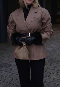 Femme portant un blazer marron avec des poignets bordés de fourrure noire, des gants noirs, et tenant un sac à main beige sur un chemin pavé de pierres.