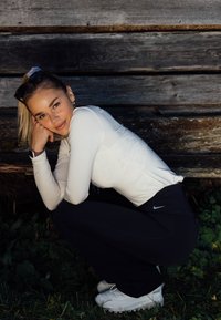 Jovem mulher agachada na relva junto a uma parede de madeira, vestindo uma camisola branca de mangas compridas, leggins pretas da Nike, ténis brancos e óculos de sol na cabeça.