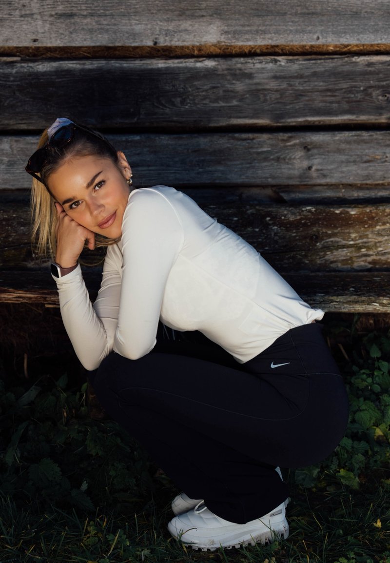 Giovane donna accovacciata sull'erba accanto a una parete di legno, indossa una maglia bianca a maniche lunghe, leggings neri Nike, scarpe da ginnastica bianche e occhiali da sole sulla testa.