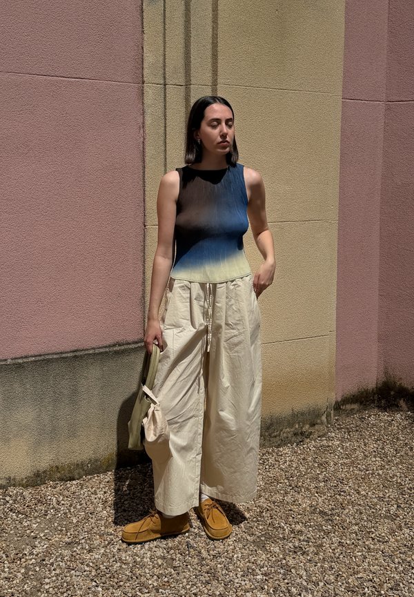 Frau mit kurzen dunklen Haaren, die ein ärmelloses Ombre-Oberteil und weite cremefarbene Hosen trägt, hält eine hellbeige Tasche und steht auf Kies vor einer strukturierten zweifarbigen Wand.
