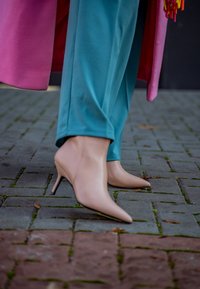 Μπεζ ψηλοτάκουνα παπούτσια με σχέδιο σε μυτερή μύτη, φορεμένα με τιρκουάζ παντελόνι σε λιθόστρωτη επιφάνεια. Ορατές λεπτομέρειες ροζ παλτού.