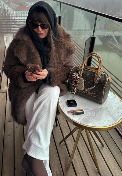 Abrigo de piel marrón, bufanda negra y gafas de sol. Pantalones de pana blancos. Botines marrones. Bolso Valentino con un colgante de leopardo. Mesa con tapa de mármol.