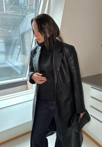 Černý kožený dvouřadý kabát, texturovaný černý rolák, tmavé džíny, držící pletenou černou kabelku. Minké přirozené světlo z okna.