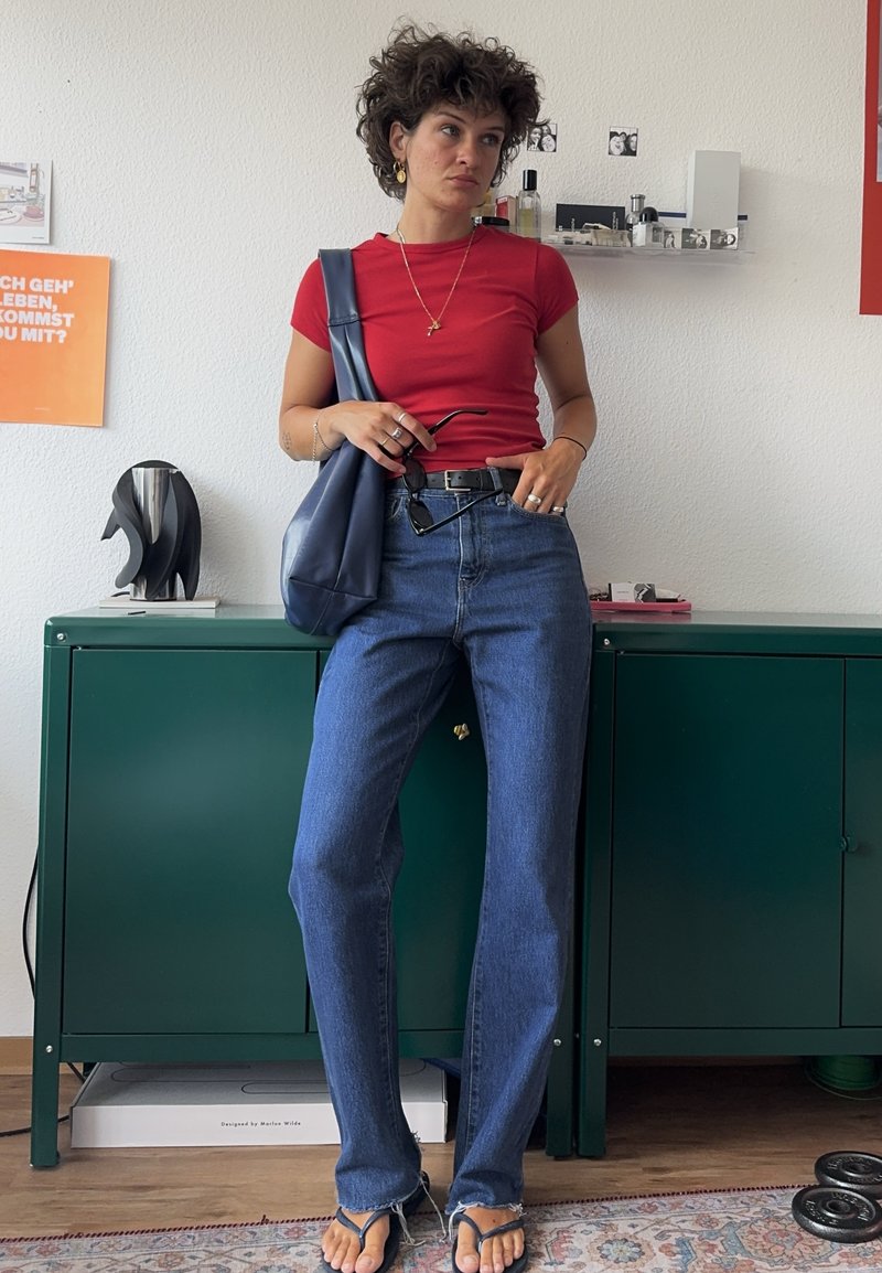 Personne aux cheveux courts et bouclés portant une chemise rouge, un jean bleu et des sandales, debout à l'intérieur tenant des lunettes de soleil et un grand sac bleu.