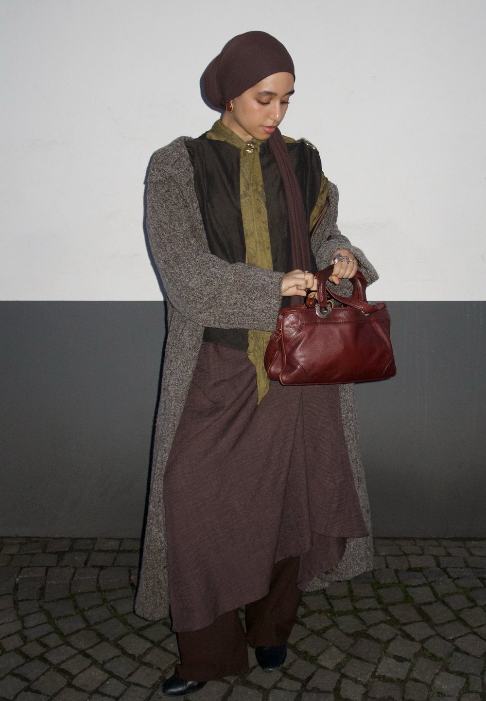Long manteau gris sur un haut foncé et une jupe marron, tenant un sac à main en cuir rouge. En arrière-plan, un mur lisse gris et blanc.