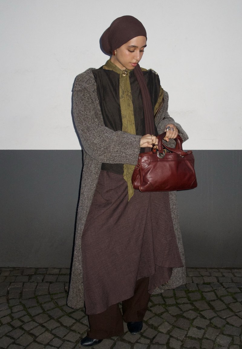 Long manteau gris sur un haut foncé et une jupe marron, tenant un sac à main en cuir rouge. En arrière-plan, un mur lisse gris et blanc.